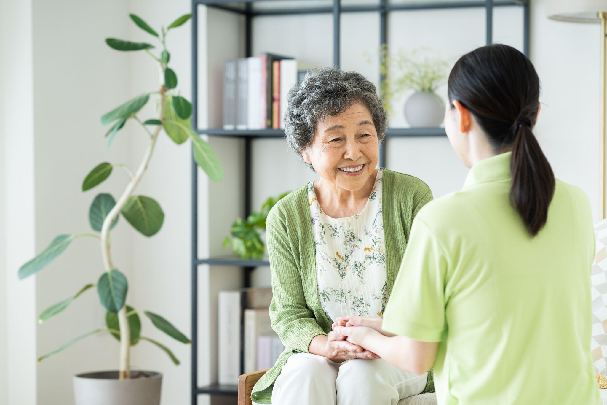 A younger adult holding the hands of an older adult, smiling at each other in a brightly lit room with plants, at a assisted living community.